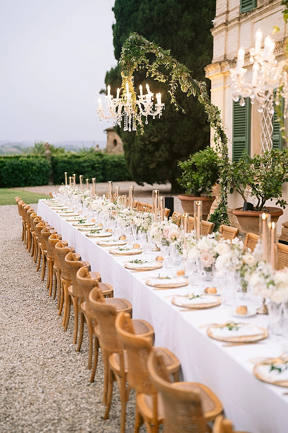 Reception tablescape with long banquet table setup on white linens, rattan chairs, florals and taper candles beneath a crystal chandelier in a villa courtyard