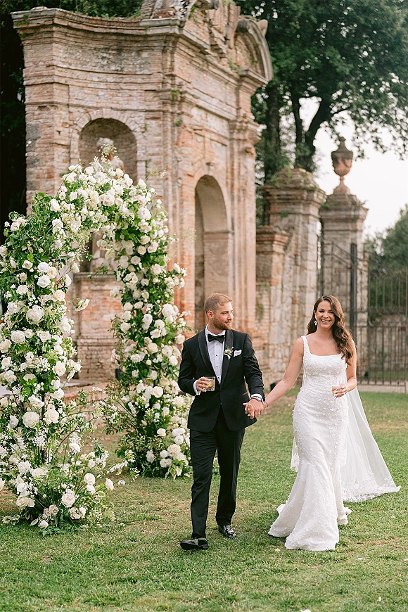 Couple portrait of bride in veil and groom in black tuxedo holding champagne glasses beneath a white floral arch at stone ruins
