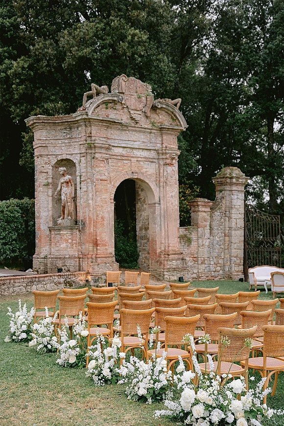 Ceremony setup with outdoor wedding ceremony chairs and white aisle flowers, framed by stone arch ruins and lush lawn greenery