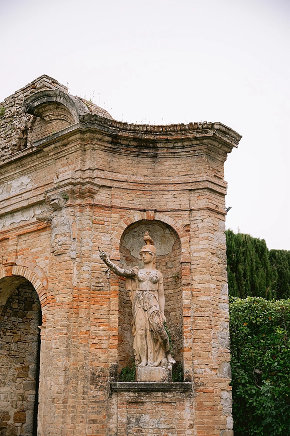 Historic venue exterior with a brick archway and stone statue set into the masonry wall, framed by hedges and trees under an overcast sky