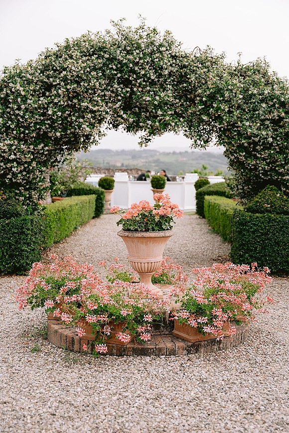 Garden ceremony arch draped in flowering greenery with potted blooms and stone urn planters along a gravel terrace path under cloudy sky