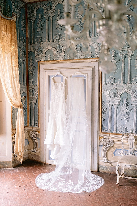 Wedding dress and veil hanging from white double doors, lace gown glowing in window light beneath a chandelier in a vintage room