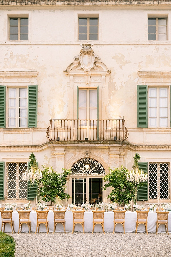 Outdoor reception tablescape with a long banquet table, white florals and candles beneath crystal chandeliers in a villa courtyard with green shutters