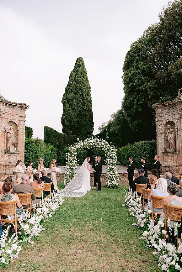 Wedding ceremony with bride and groom at a round white floral arch, long veil flowing down a flower-lined aisle on a garden lawn