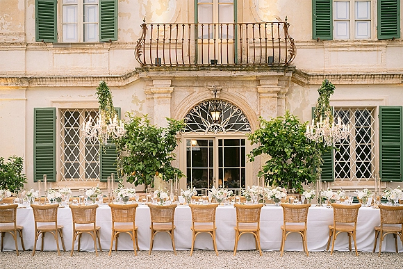 Reception tablescape for an outdoor wedding reception with a long white-clothed banquet table, taper candles, florals, chandeliers in a villa courtyard