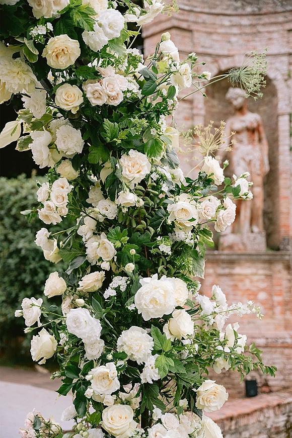 Wedding floral arch of white roses and greenery on an asymmetrical frame, set against a stone building and brick wall in a garden walkway