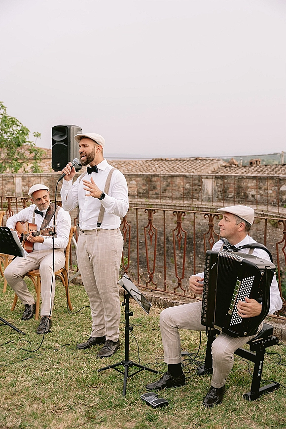 Wedding band playing live wedding music outdoors, singer at microphone with guitar and accordion beside a stone wall and iron railing