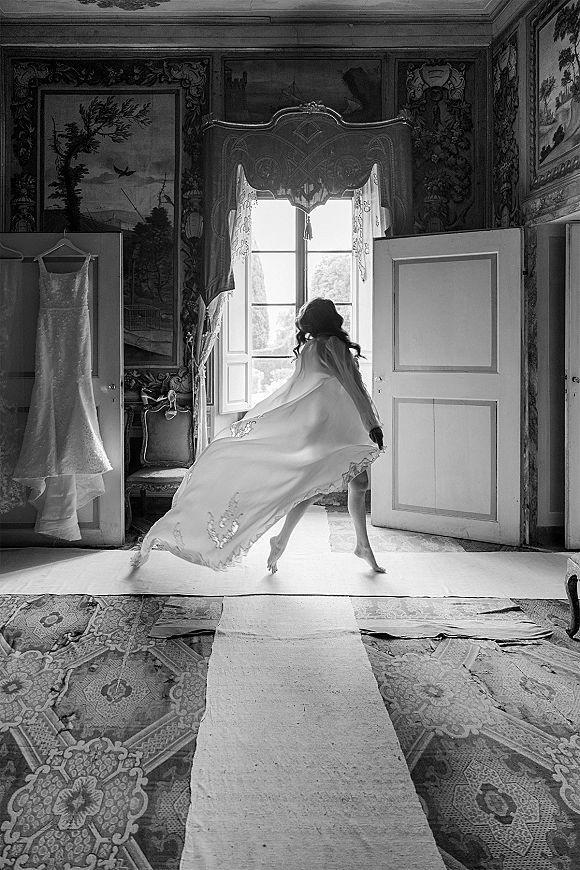 Bridal portrait in black and white of a bride in robe with lace trim by a tall window, ornate mural wallpaper behind her