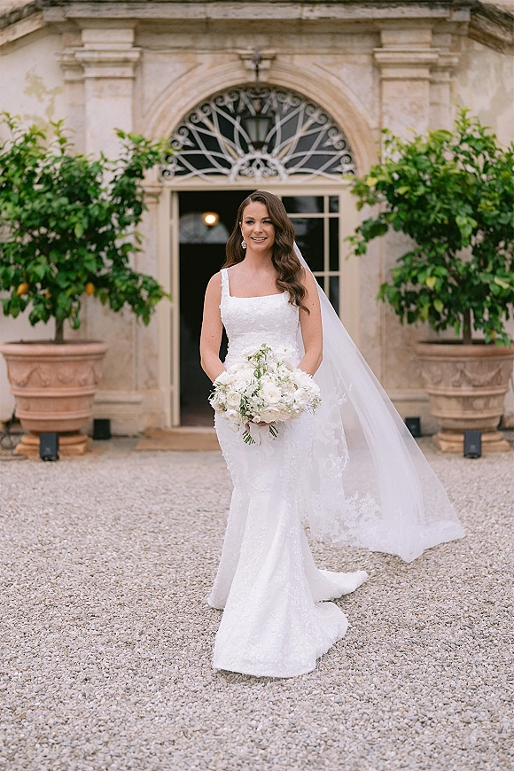 Bridal portrait of a bride holding bouquet of white roses and greenery, wearing a lace gown and cathedral veil in a stone courtyard doorway