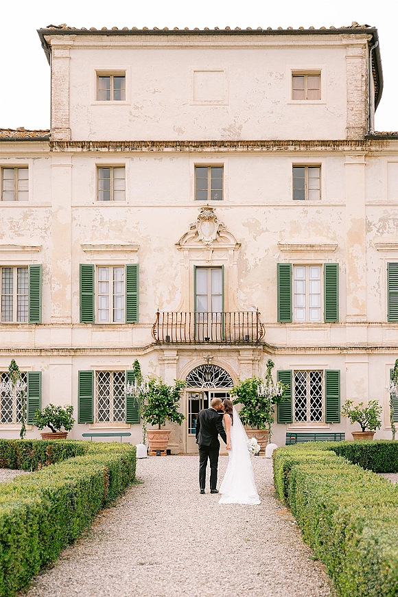 Wedding couple portrait of bride and groom kissing, her veil train and bouquet flowing before a villa with green shutters and balcony