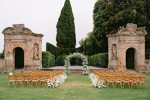 Ceremony setup with a white floral arch and aisle florals, wooden chairs on a grass lawn framed by hedges and stone archways