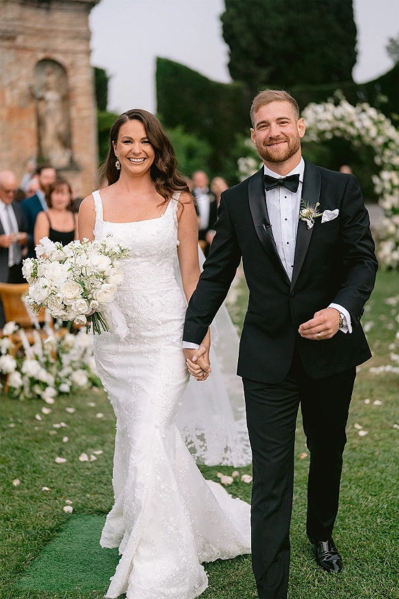 Wedding recessional as bride and groom walk the aisle holding hands, bride with white bouquet and long veil on garden lawn with guests