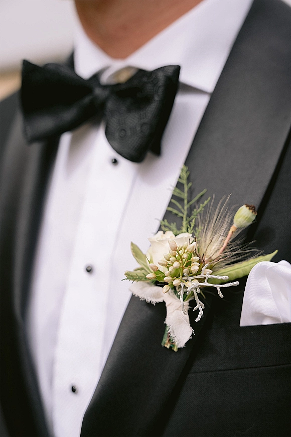 Groom boutonniere with a white rose boutonniere and greenery pinned to a black tuxedo lapel beside a black bow tie, blurred background