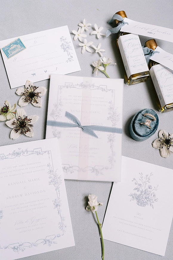 Invitation suite wedding invitation flat lay with blue and white cards, vellum overlay, ribbon, ring box and mini favor bottles on gray fabric