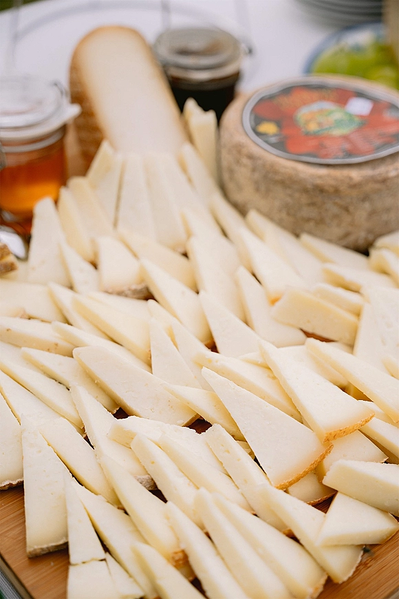 Cheese board with sliced cheese, a cheese wheel, baguette, and jam jars arranged on a wooden board on a white tablecloth