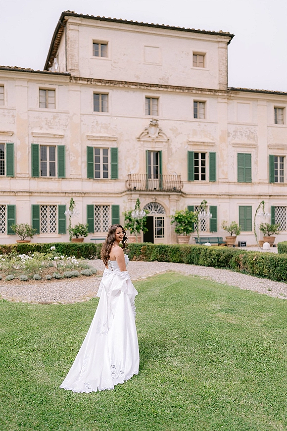 Bridal portrait of a bride looking back in a strapless wedding dress with satin overskirt and lace appliqué before a villa with green shutters