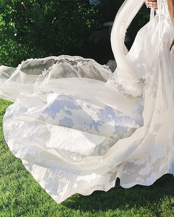 Wedding dress detail with a wedding dress train swirling in sunlight, lace and floral appliqué visible on tulle over a green lawn