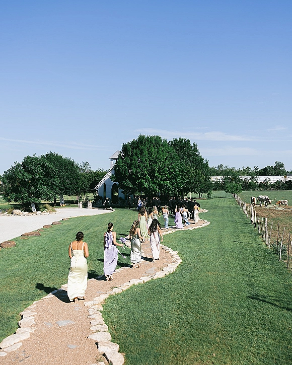 Wedding processional with bridesmaids walking aisle in pastel dresses along a gravel path toward a white chapel, horses in the pasture beyond