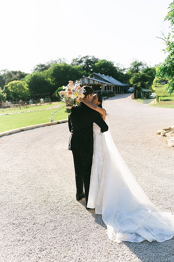Wedding kiss as bride and groom embrace on a gravel road by a barn, her veil and long train flowing with bouquet over shoulder