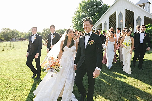Wedding recessional as bride and groom walking hand in hand past cheering guests on a sunny chapel lawn, bouquet and veil flowing