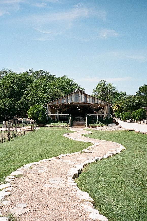 Outdoor reception venue with barn wedding venue stage and string lights accent, set on a green lawn beside a gravel path under blue sky