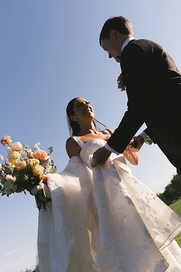 Couple portrait of bride and groom holding hands, bride gazing up at him, with bouquet and veil in a grassy field under blue sky