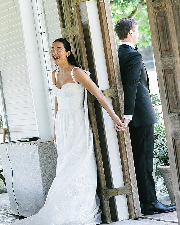 First look moment as bride and groom hold hands around a corner by wooden porch doors, her earrings catching light, both smiling