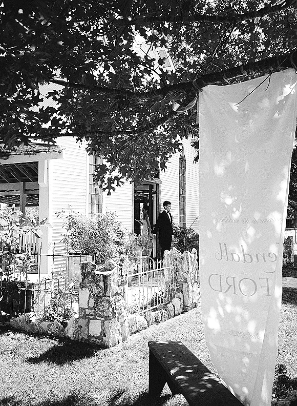 Wedding venue exterior with a fabric flag banner and wrought iron gate on a stone wall, facing a white chapel under trees in dappled shade