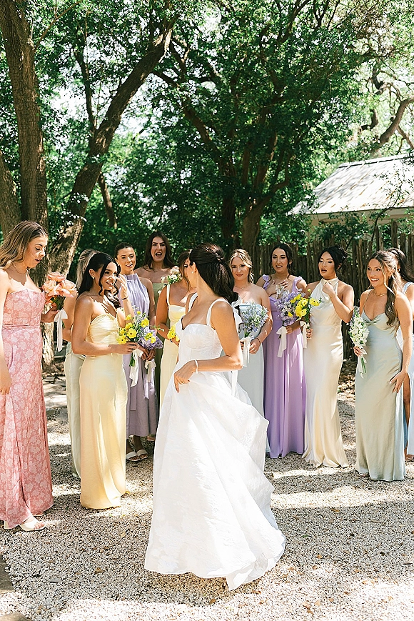 Bridesmaid reveal as the bridal party gathers around the bride in a strapless wedding dress, holding bouquets on a gravel path by trees