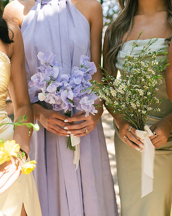Bridesmaid bouquets of sweet peas with white filler flowers, held by bridesmaids in lavender, sage, and yellow dresses in dappled greenery