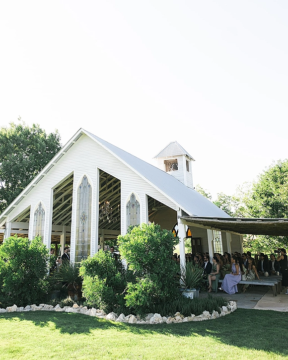 Outdoor wedding ceremony at an open air chapel, with guests on wooden benches under canopy shade beside a white bell tower