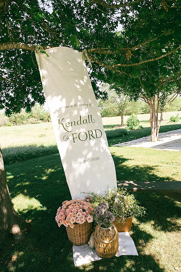 Wedding welcome sign in cream fabric with calligraphy, tied ribbons, and rose florals in wicker baskets under a tree canopy on lawn