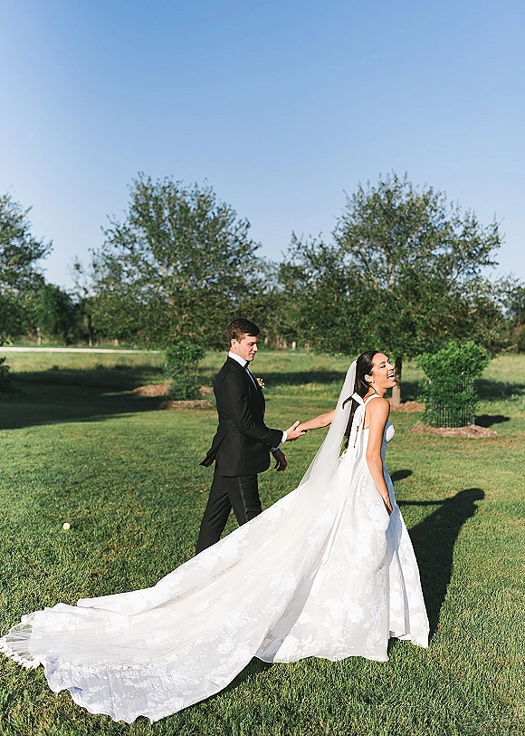 Couple portrait of bride and groom holding hands on a grassy lawn, bride looking back with veil and long train beside groom in tuxedo