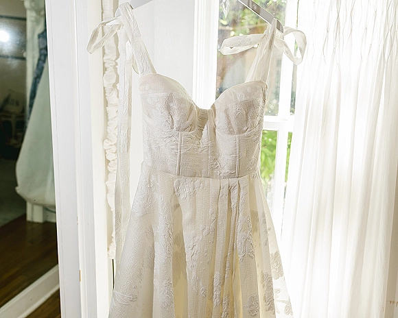 Wedding dress hanging on a hanger, strapless wedding dress with corset bodice and bow shoulder ties by a window with sheer curtains