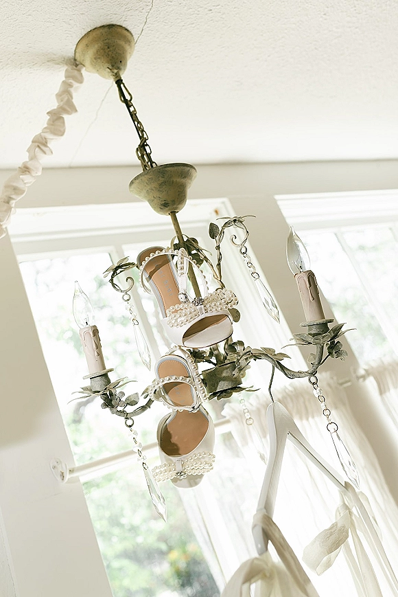 Bridal shoes with pearl wedding heels hang from a crystal chandelier, sparkling in daylight by large windows with sheer curtains