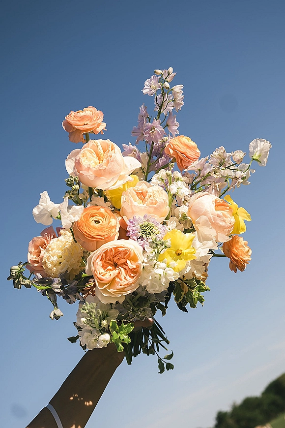Wedding bouquet with hand-tied stems of peach roses, ranunculus, yellow and white blooms with greenery, held up against blue sky