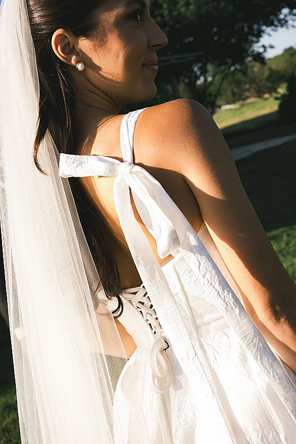 Bridal portrait of a bride in profile wearing a long veil and pearl stud earrings, with bow-strap white dress in sunlit greenery outdoors
