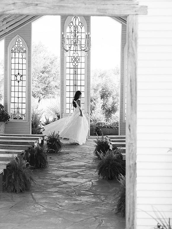 Bridal portrait of a bride twirling her tulle wedding dress down a stone chapel aisle, chandelier overhead and stained glass windows behind