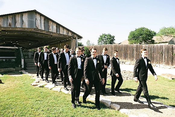 Groomsmen portrait of men in black tuxedos with boutonnieres, walking along a gravel path beside a rustic barn under blue sky