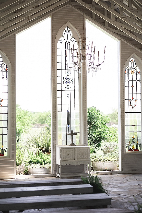 Chapel ceremony setup with stained glass windows, white altar table and cross beneath a chandelier, open wall framing garden greenery