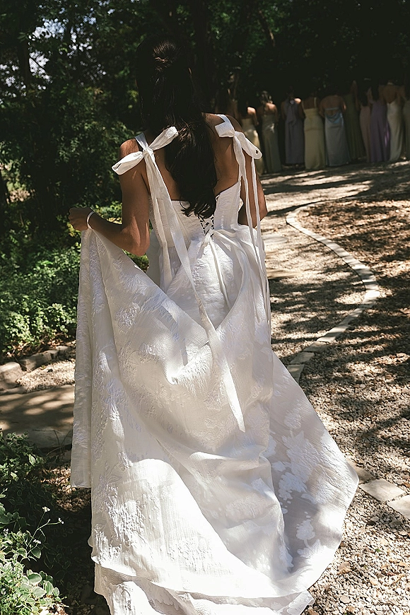 Bride walking away in a ribbon-strap wedding dress back view, holding her long train on a gravel garden path with pastel bridesmaids behind