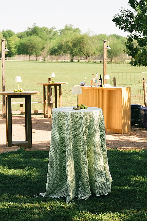 Outdoor wedding bar with portable wedding bar setup, liquor bottles, limes, glasses and lamp-lit cocktail tables on a grassy lawn by trees