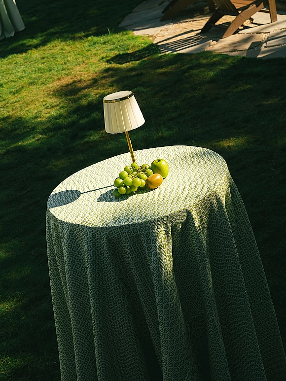 Cocktail table decor with a green patterned tablecloth, small lamp centerpiece, and fruit accents on a sunlit patio lawn setting
