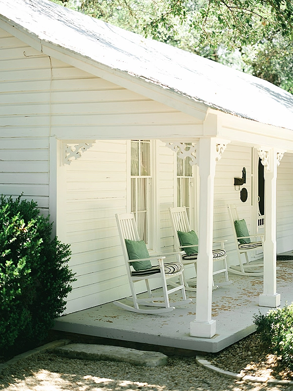 Wedding venue porch with white rocking chairs and striped cushions on a white cottage exterior, shaded by shrubs beside a gravel driveway