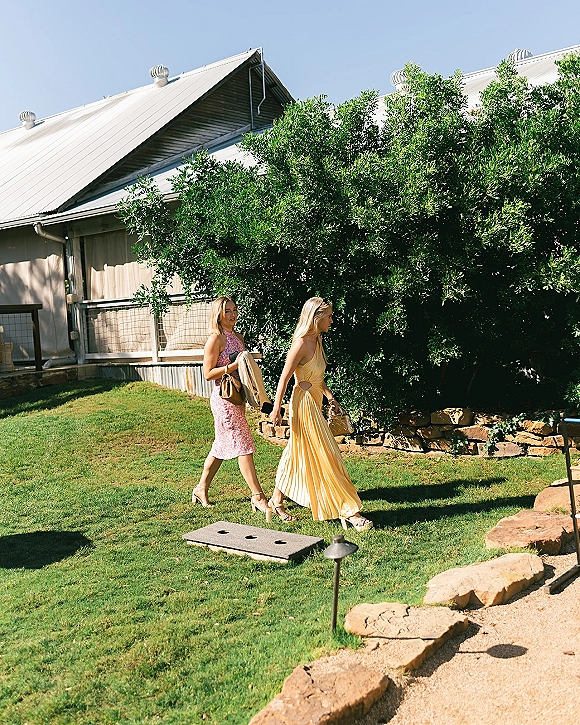 Wedding guests in wedding guest dresses walk along a garden path, one in a yellow pleated maxi dress under blue sky by a farmhouse