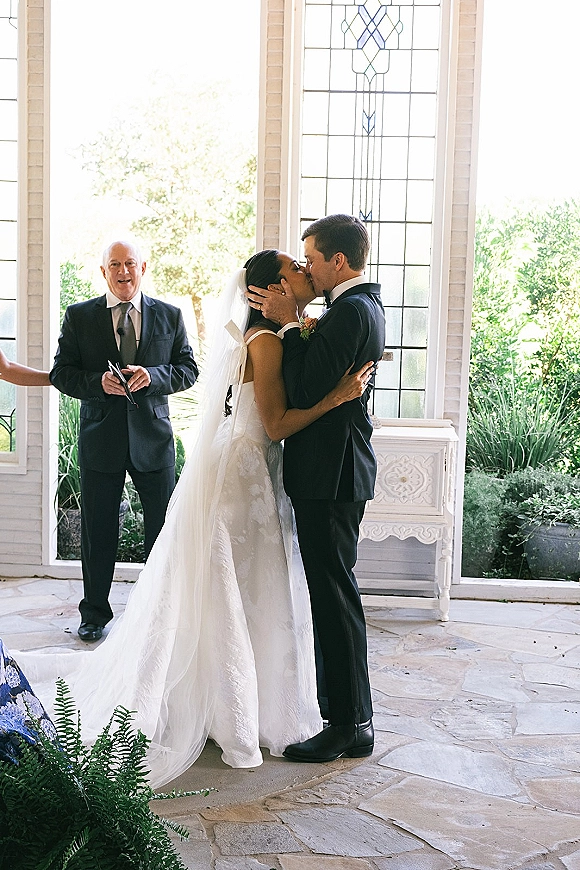 Wedding kiss as bride in strapless gown and long veil kisses groom in black suit before stained glass, white brick, and greenery backdrop