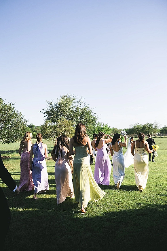 Wedding processional of bridesmaids walking aisle ahead of the bride with long veil and bouquet across a sunny grassy field with trees