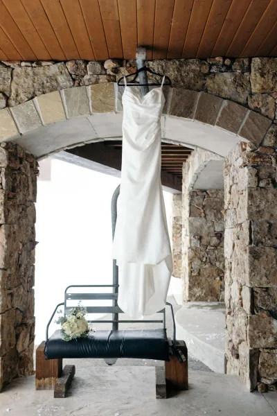 Wedding dress hanging on a dress hanger, strapless satin gown with a bridal bouquet beside it under a stone archway outdoors