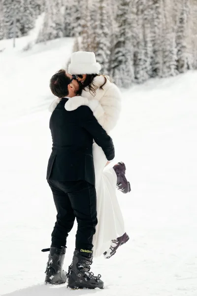 Wedding kiss portrait of groom lifting bride in a snowy wedding portrait, her faux fur jacket and hat framed by pine trees in winter forest