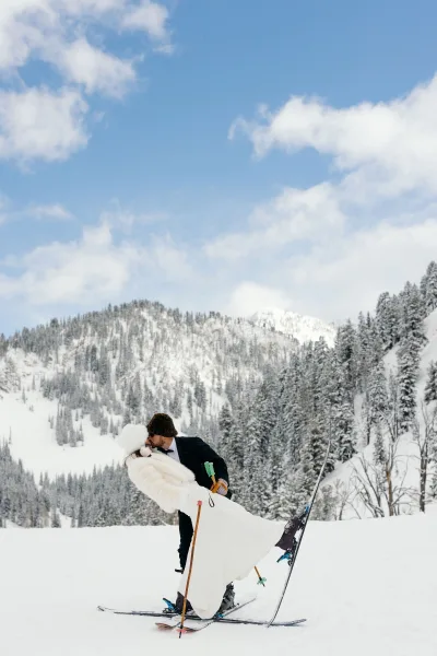 Wedding kiss portrait of bride and groom on skis in the snow, holding poles under pine trees with mountain peaks and blue sky behind